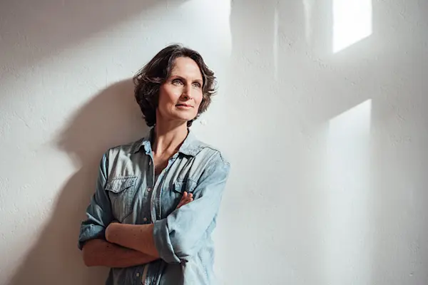 Woman in denim shirt with arms folded standing next to a window casting shadows. AW508 