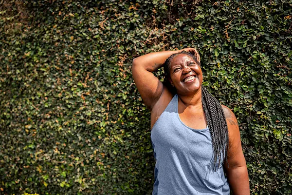 Middle aged Black woman in workout clothing, her hair in braids, smiling with her hand resting on her head in front of a foliage covered wall. thumbnail
