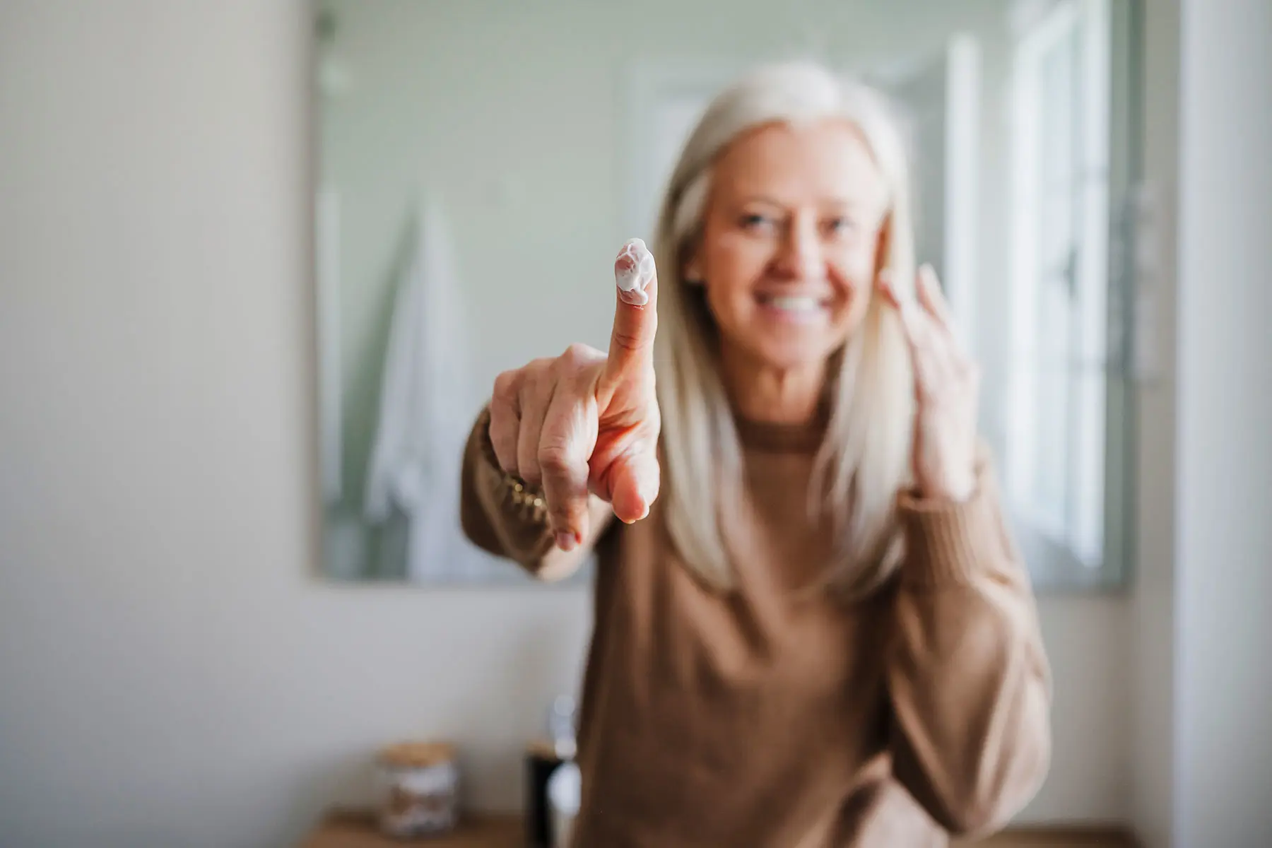 Woman with shoulder length white hair in bathroom smiling with face out of focus, hand in focus with moisturizer on finger. AW552