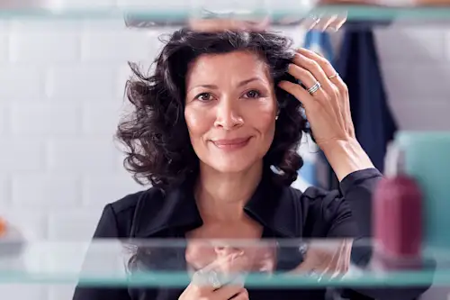 Latina woman in reflection of medicine cabinet combing her hair back with her fingers, smiling. AW395