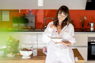 Woman in kitchen in pajamas sprinking berries on her bowl of yogurt (thumbnail)