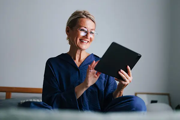 Woman in bed wearing blue pajamas and glasses, smiling using tablet. thumbnail