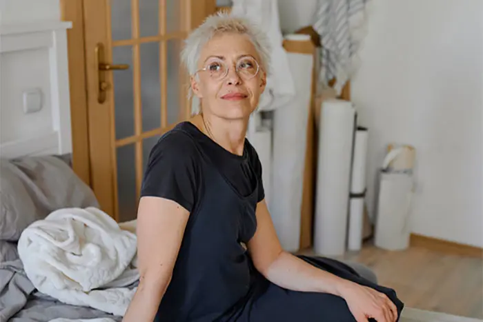 Woman with short blonde hair in glasses sitting on unmade bed looking away. AW401