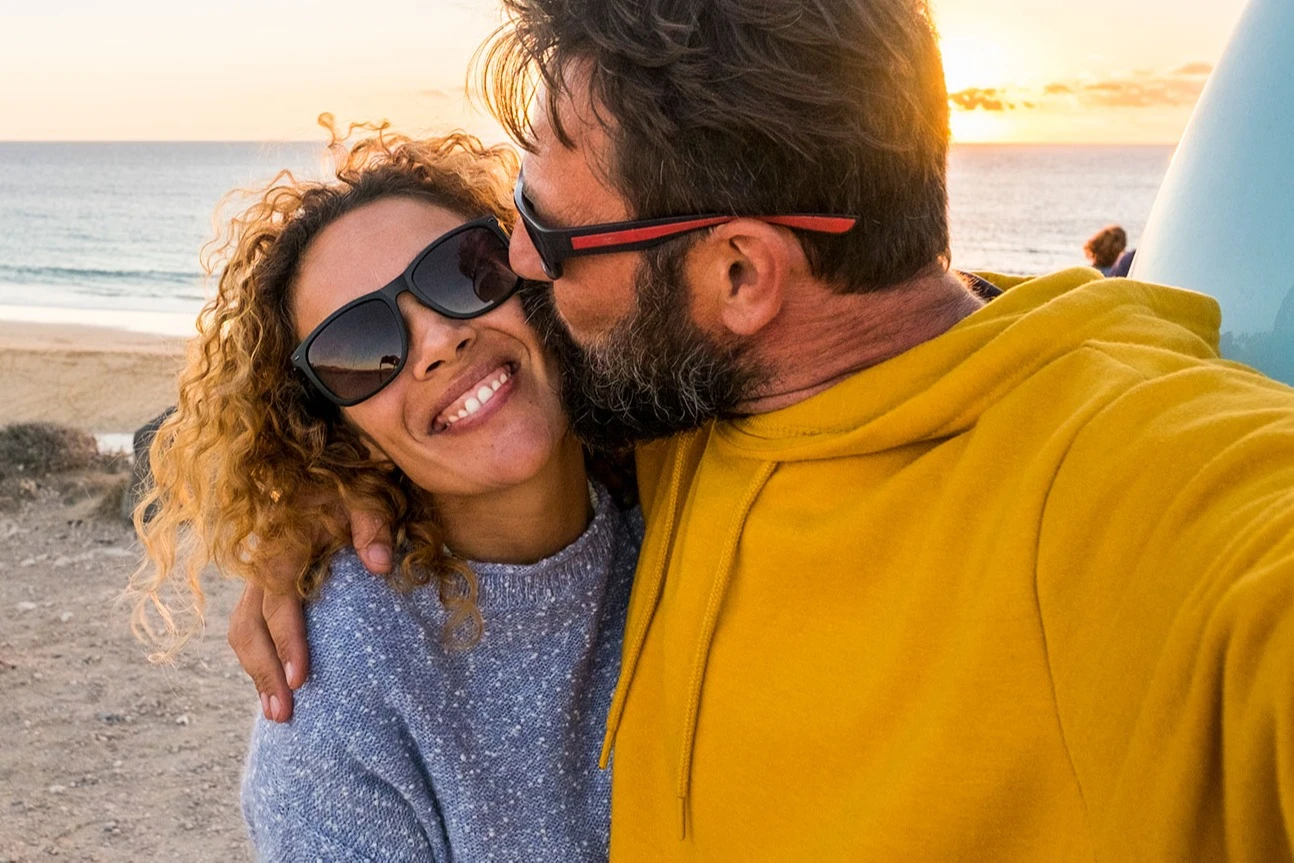 Woman smiling in sunglasses being kissed on the cheek by bearded man at beach. AW323