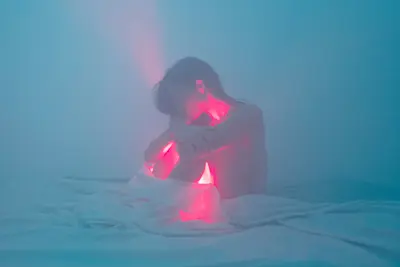 Woman sitting on a bed in a blue, dimly lit room, with a red light glowing from sheets under her.  (thumbnail)