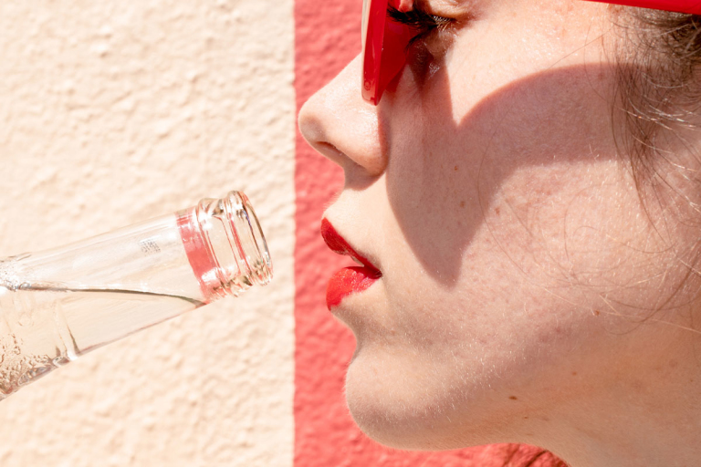 Closeup on the side of a face. A bottle of water is being lifted toward a mouth wearing red lipstick.