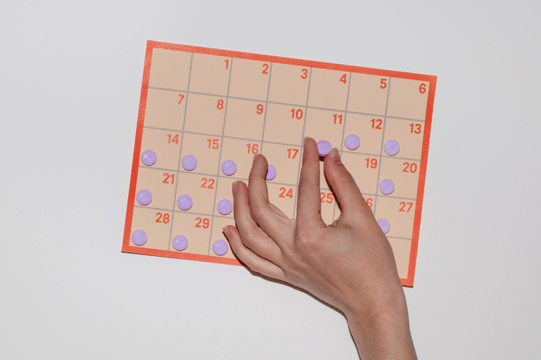 Hand holding purple pill against an orange and peach calendar with other purple pills against a gray neutral background