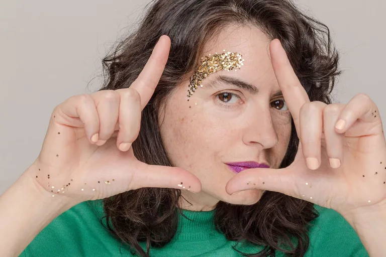 Woman with black hair and purple lipstick in a green shirt making Ls with hands with glitter on temple against a neutral gray background