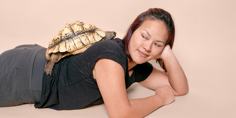 A woman laying on her stomach poses with a turtle on her back. 
