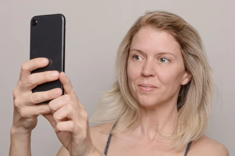 Woman with blonde hair holding up phone and looking away from the camera against a neutral gray background