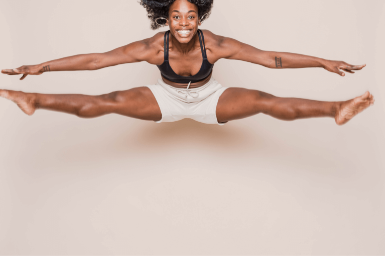Woman in a sports bra and shorts jumping into a split against a light neutral background