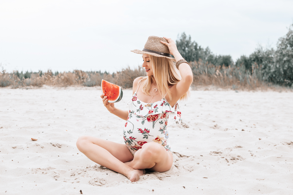 Young happy pregnant woman in a swimsuit and hat in the sand