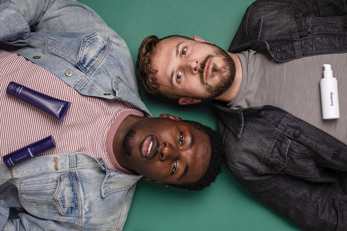 Two men are laying down, their bodies pointing in opposite directions. Curology bottles rest on their chest.