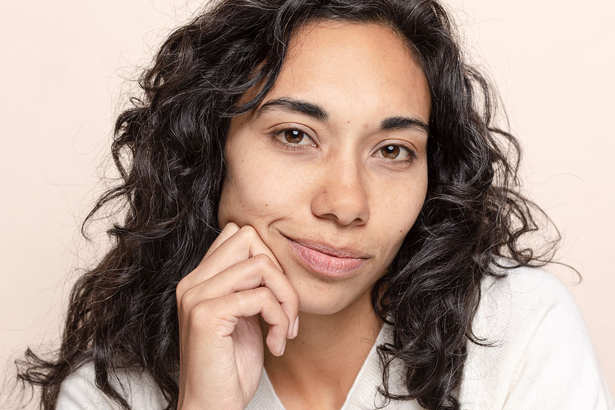Woman smiling without teeth with hand near face in front of light neutral background
