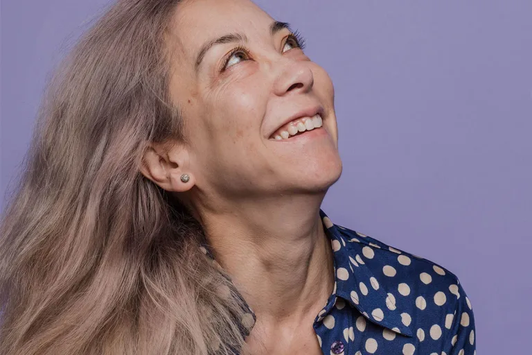Woman with gray hair in a dotted shirt smiling and looking away from camera against a purple background