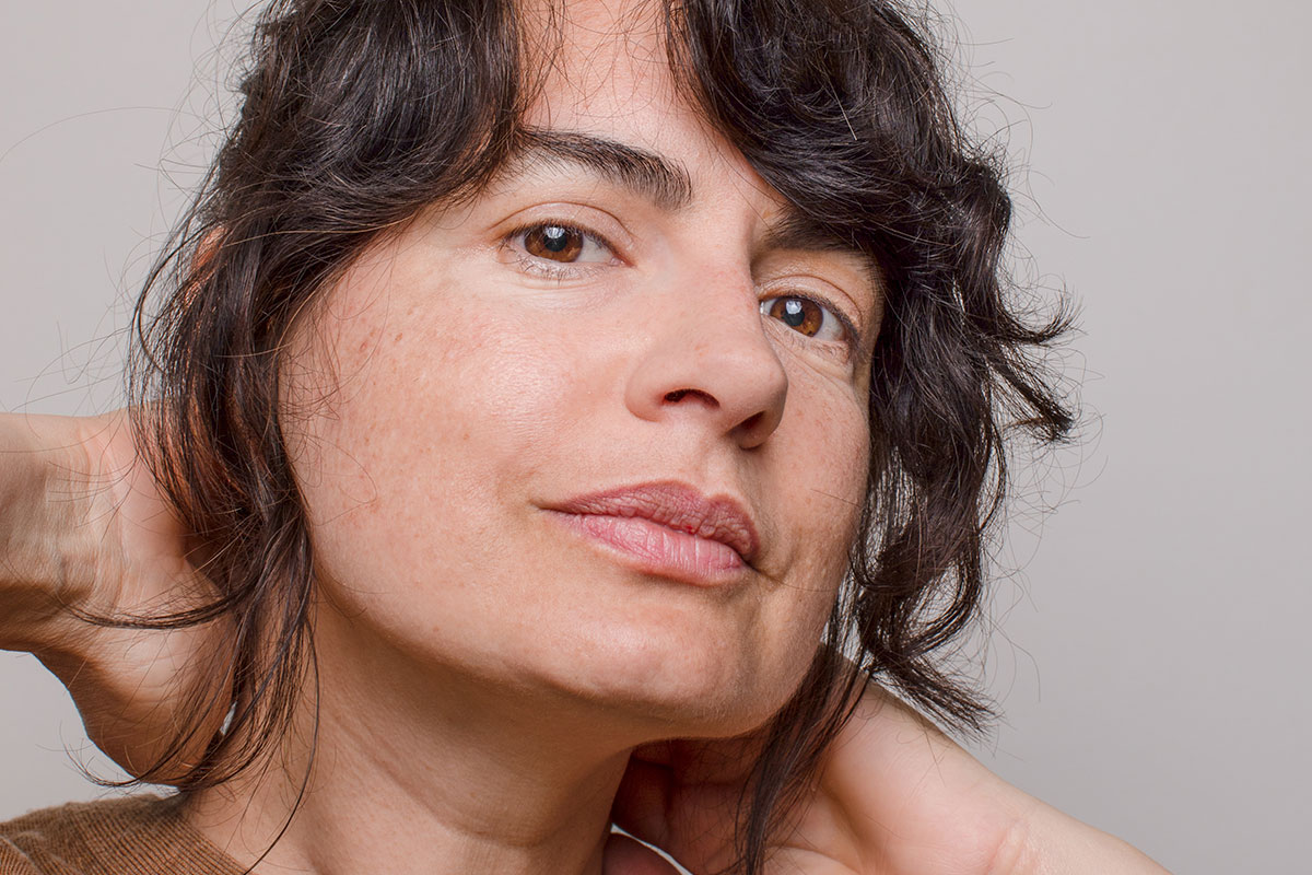 Closeup of a woman's face. Her hands are resting on the back of her neck and she is looking into the camera.