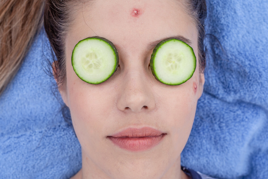Close-up of a woman's face. She is laying on a blue towel with cucumbers resting on her eyes.