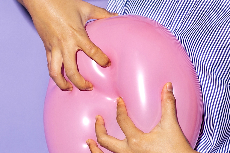 Closeup of hands pressing into a pink balloons, with a pinstriped shirt visible, all against a purple background