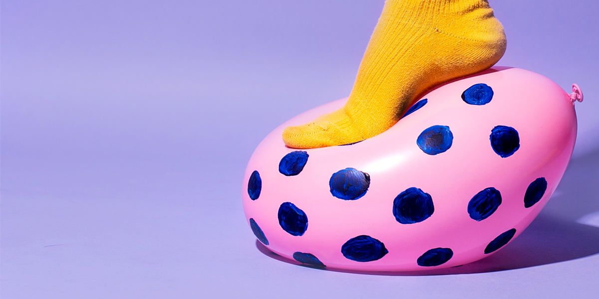 Closeup of foot in a yellow sock stepping and pressing a pink balloon with blue dots, all against a purple background