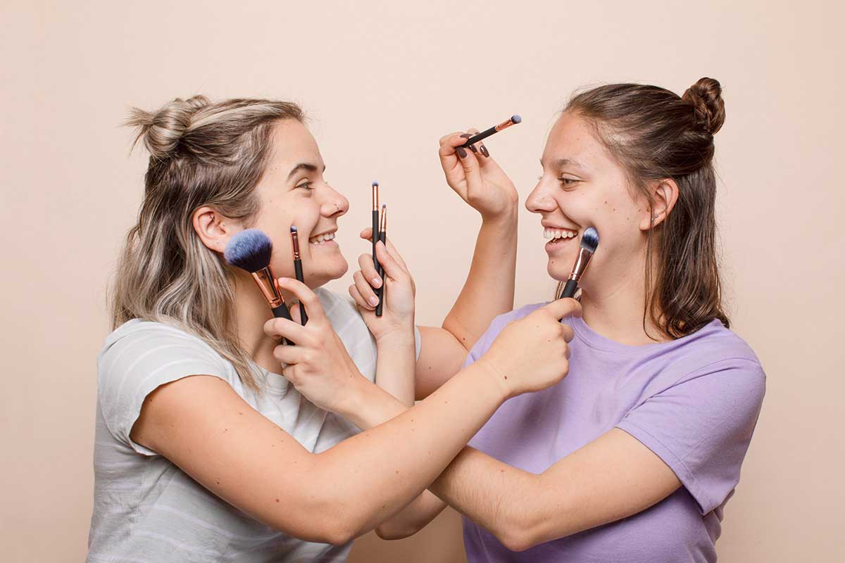 Two women facing one another and laughing, holding makeup tools and applying makeup to one another's faces against a light peach background