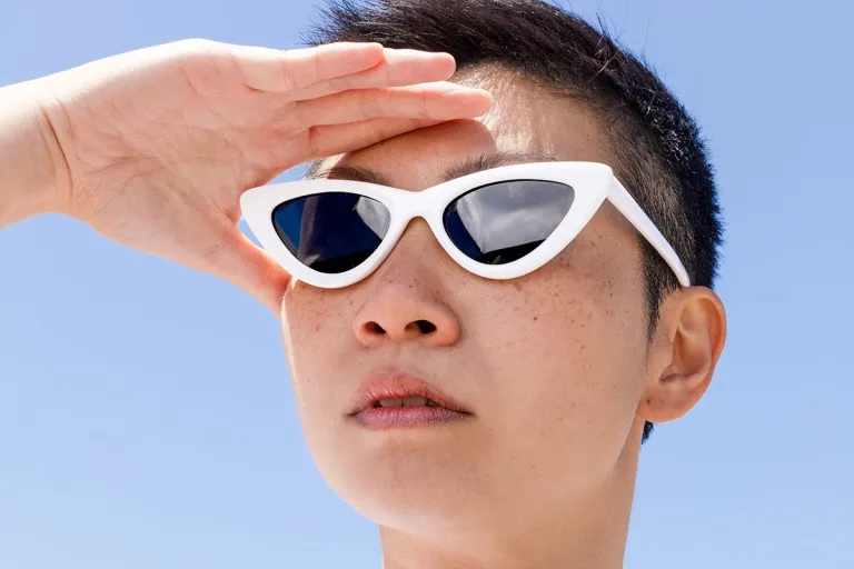 Woman with short black hair in white sunglasses with hand shading forehead against a blue sky background