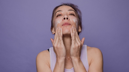 Woman rubbing skincare cream into her face with purple background