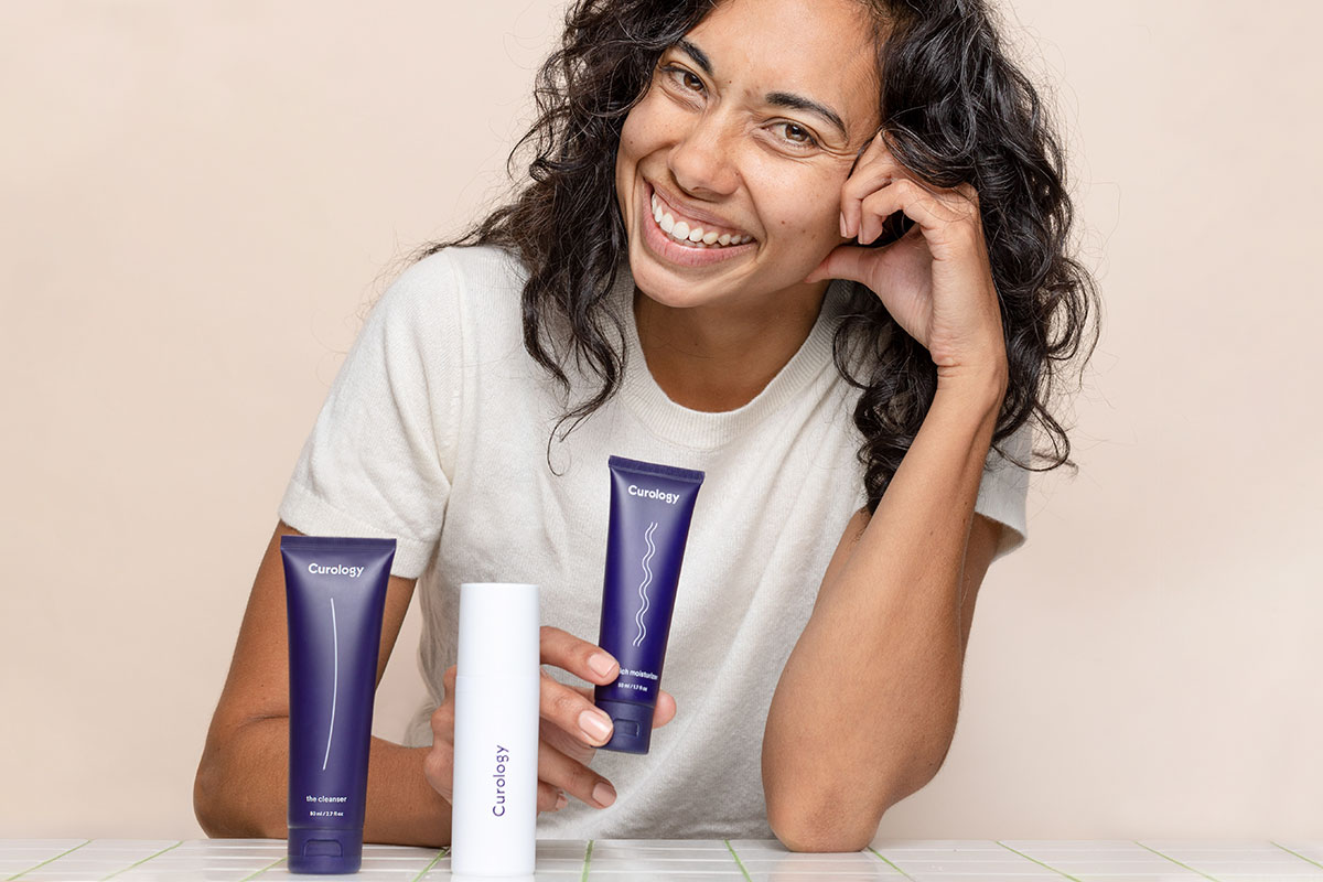 A woman smiling and resting her head in her hand, posing with the Curology set. 