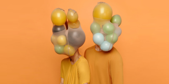Two people in yellow and orange shirts with yellow, green, and blue balloons in place of heads, all against an orange background