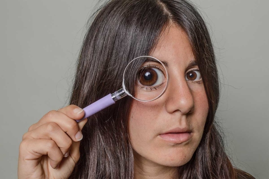 Portrait of a woman holding a magnifying glass in front of her eye so that it looks comically large 