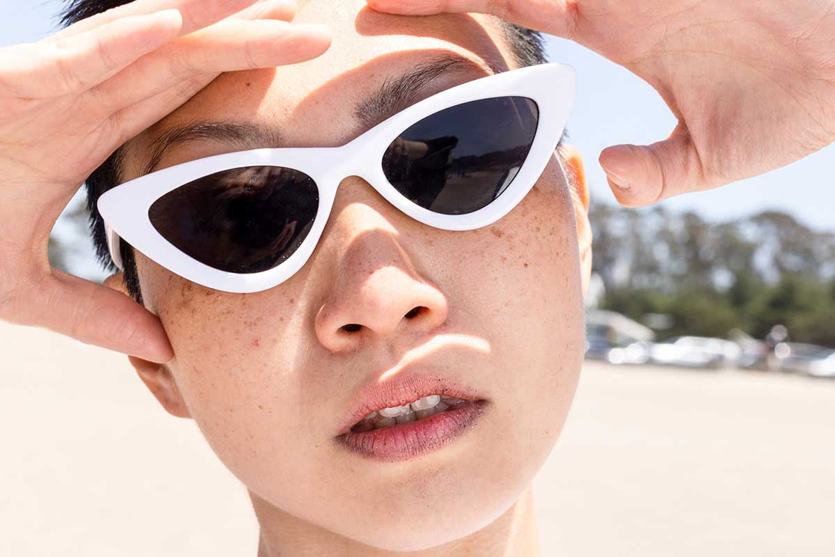 Woman in white sunglasses with hands by forehead against an outdoor beach
