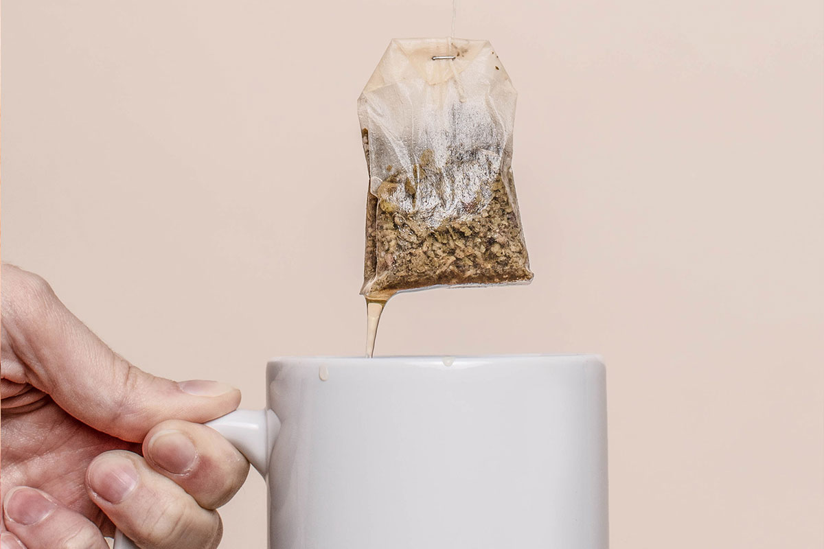 Closeup of hand holding white mug below tea bag against a neutral background