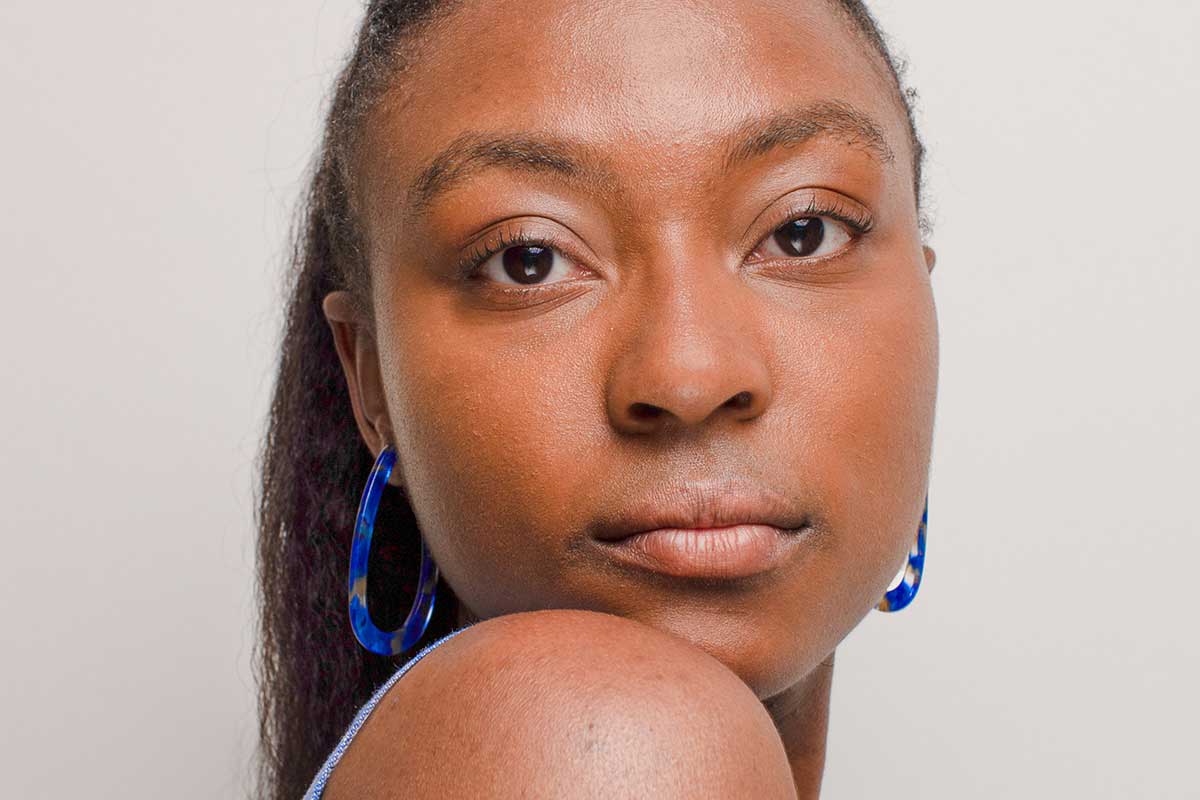 Closeup of woman with ponytail and blue hoop earrings looking at camera over right shoulder against a light gray neutral background