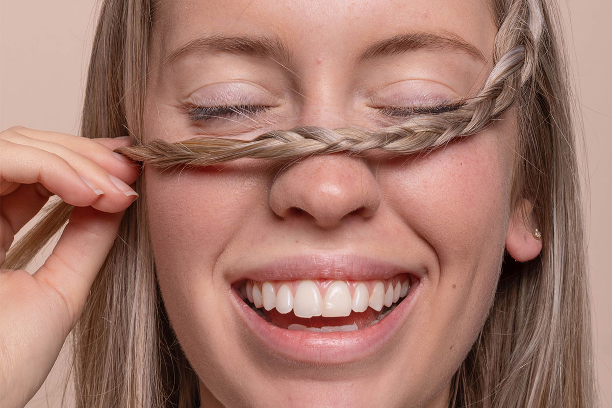 Woman with blonde hair smiling with eyes closed and holding braid over face in front of a neutral background