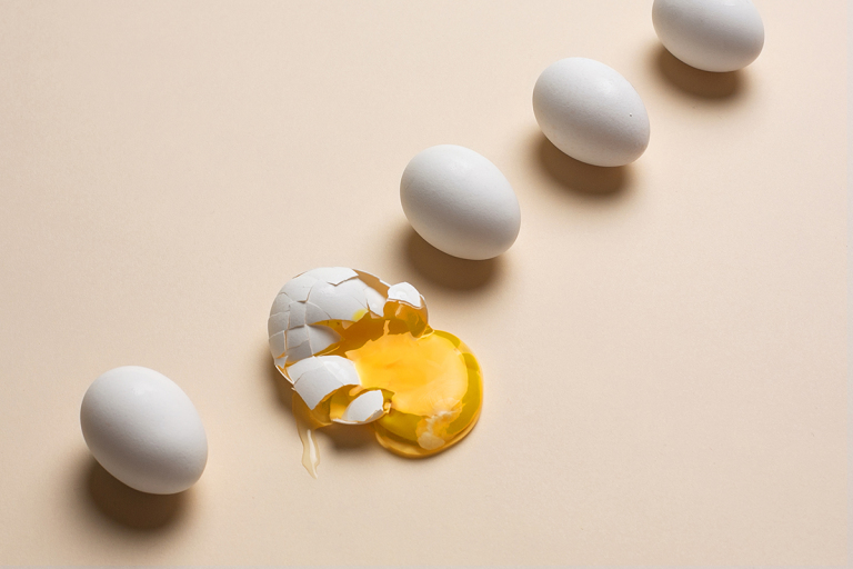 Diagonal row of 5 eggs with the second egg to the left cracked and orange yolk spilling out, all against a light neutral background