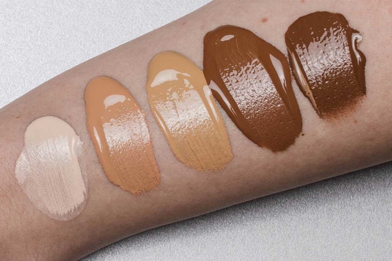 Closeup of five shades of foundation swatches on an arm against light gray neutral background