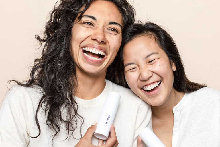 Two women laughing and leaning on each other while holding bottles of Curology custom cream