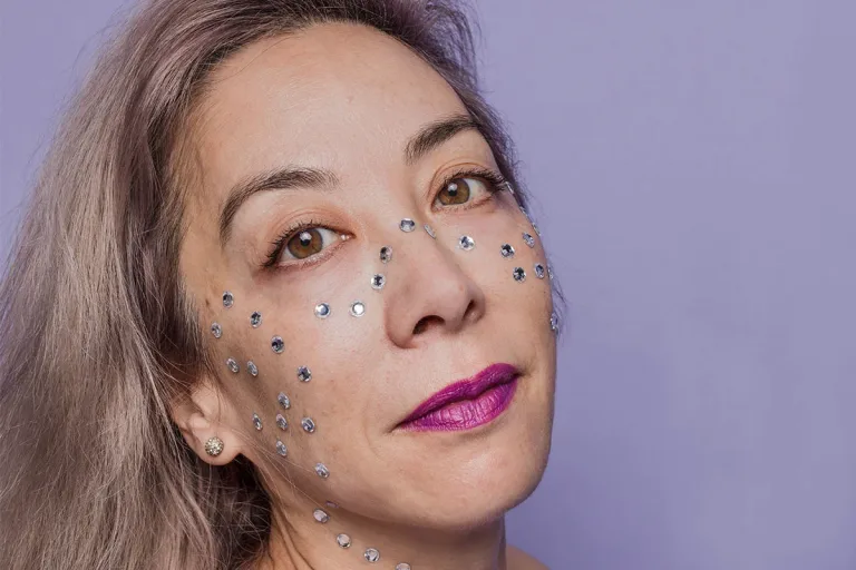 Woman with gray hair with purple lipstick and silver rhinestones on face against a purple background