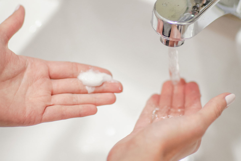 Closeup of hands with white nail polish holding  cream or soap under a faucet of running water
