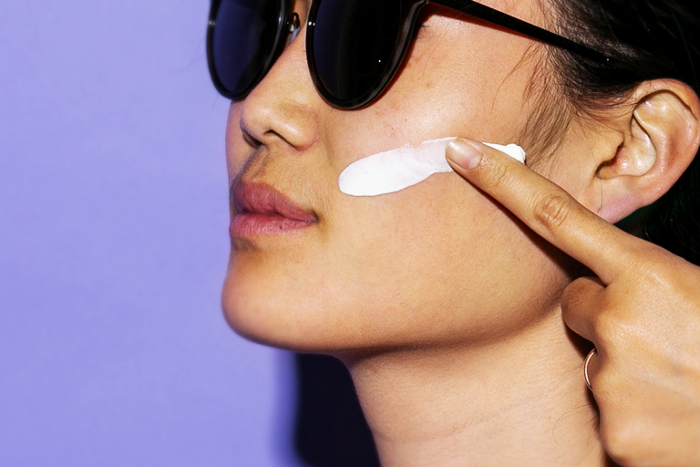 Closeup of woman in sunglasses facing left and applying cream to left cheek with index finger against a purple background