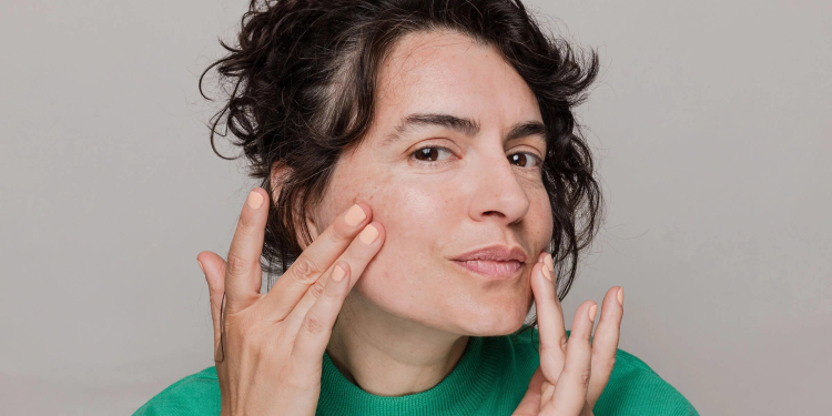 Woman with black hair in green shirt touching face with hands against a neutral gray background