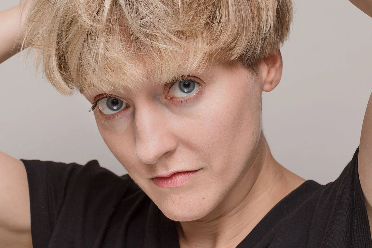 Woman with blonde hair in black shirt looking up at camera against a neutral gray background