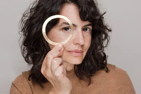 Woman with black hair in a brown shirt holding a palm-sized metallic round ring near face against a neutral gray background