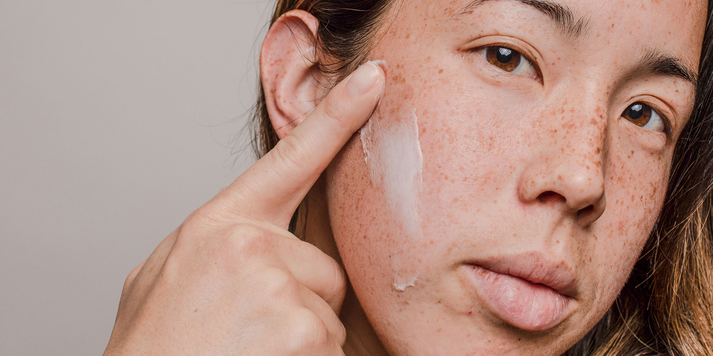 A close-up of a person's face, smearing cream across their cheek with one finger
