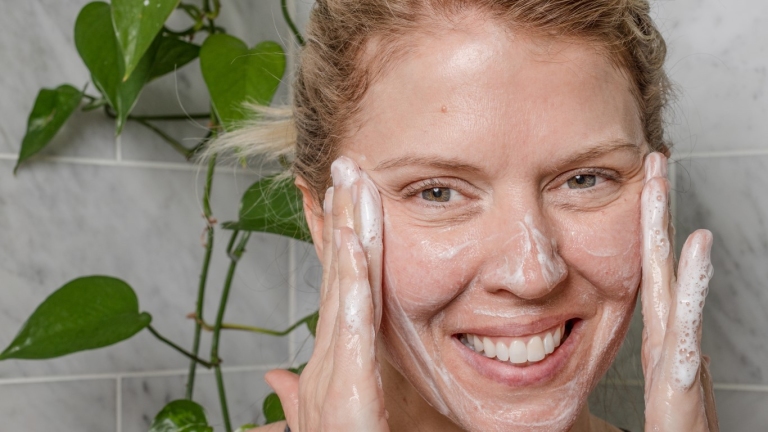 Woman smiling and applying cleanser to face in front of a plant and gray tile background