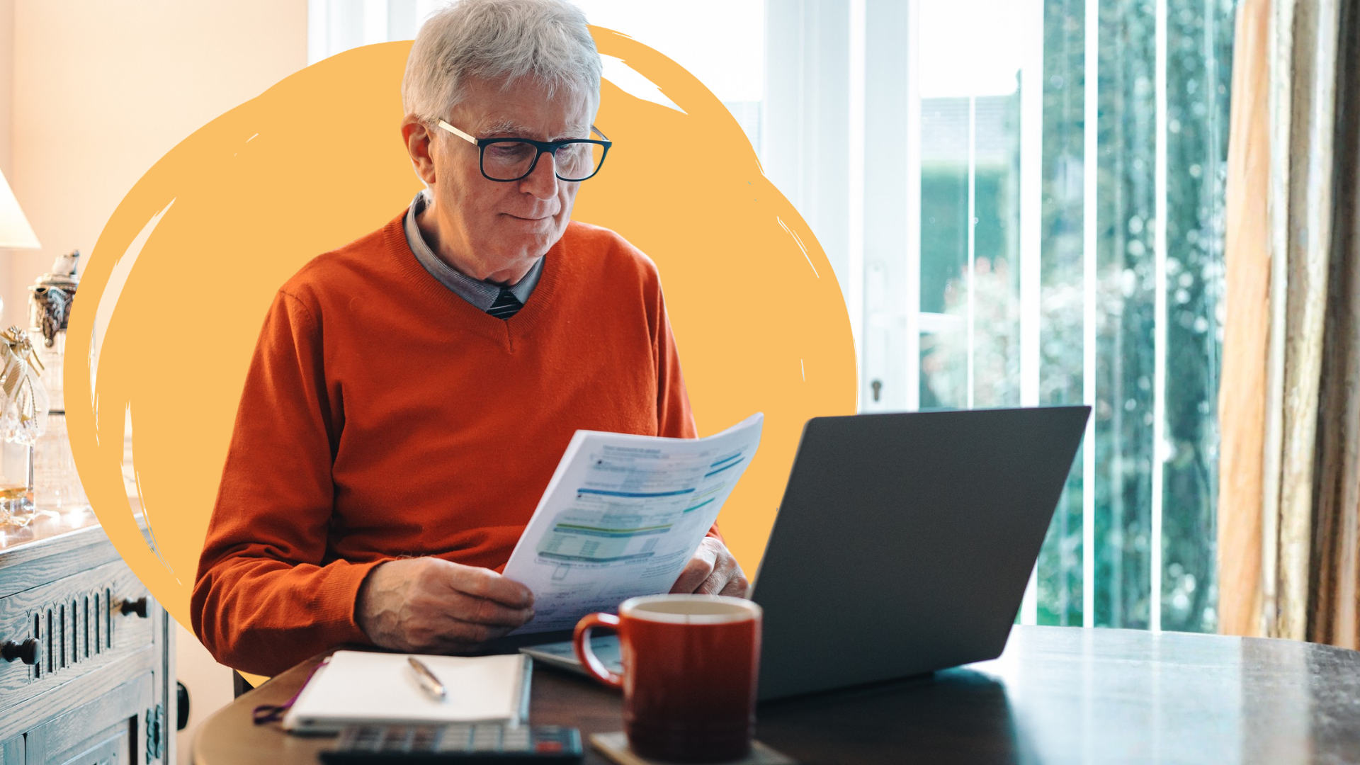 A elderly man wearing glasses sits at a table holding energy bills. He has a laptop in front of him.