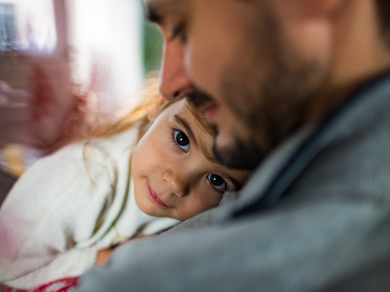 Young girl looking at camera with small smile on her face, leaning on her fathers chest who is looking down at her smiling