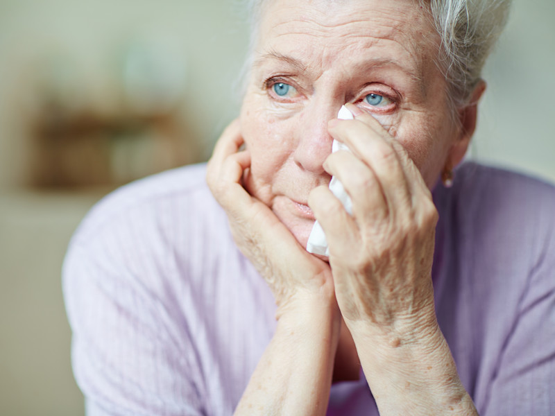 An older woman looks visibly upset and holds a handkerchief to her eye.