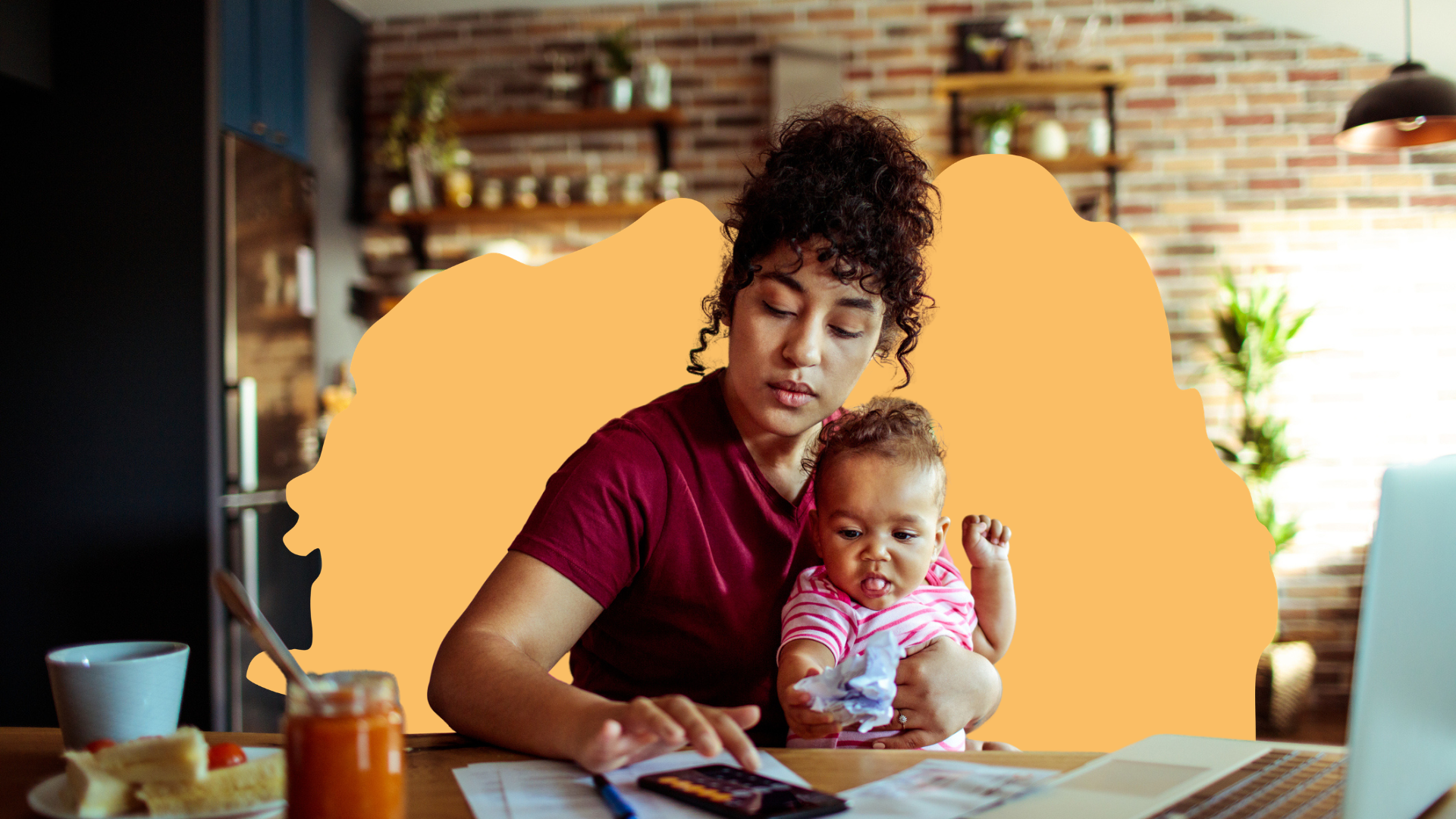 Image shows woman sitting at a table with her baby on her lap. She is using a calculator on her phone, and in front of her is a laptop and some paperwork.