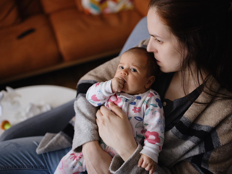 A mother is sitting on a chair, hugging her baby on her lap. She looks anxious.