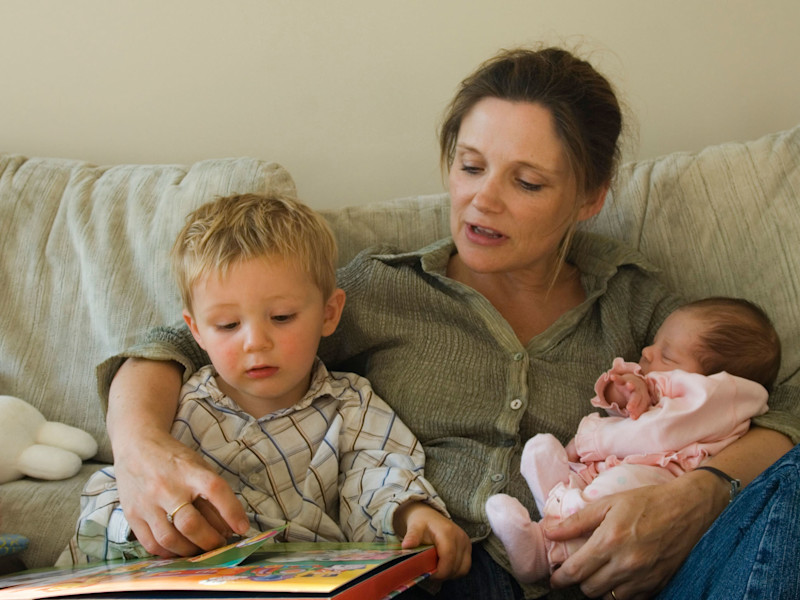 A mother sits on a sofa with a young boy and a baby in her arms. She is reading to the young boy.